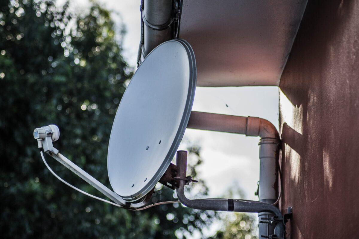 Closeup shot of a white satellite dish on fixed on a brown wall with trees on the background